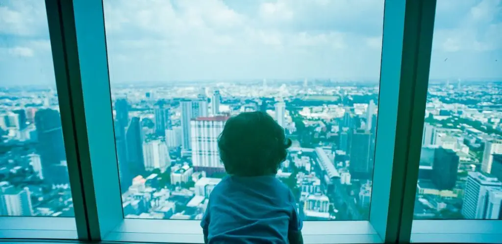 A young child looks out over the Bangkok skyline through a large window, capturing a panoramic cityscape view.